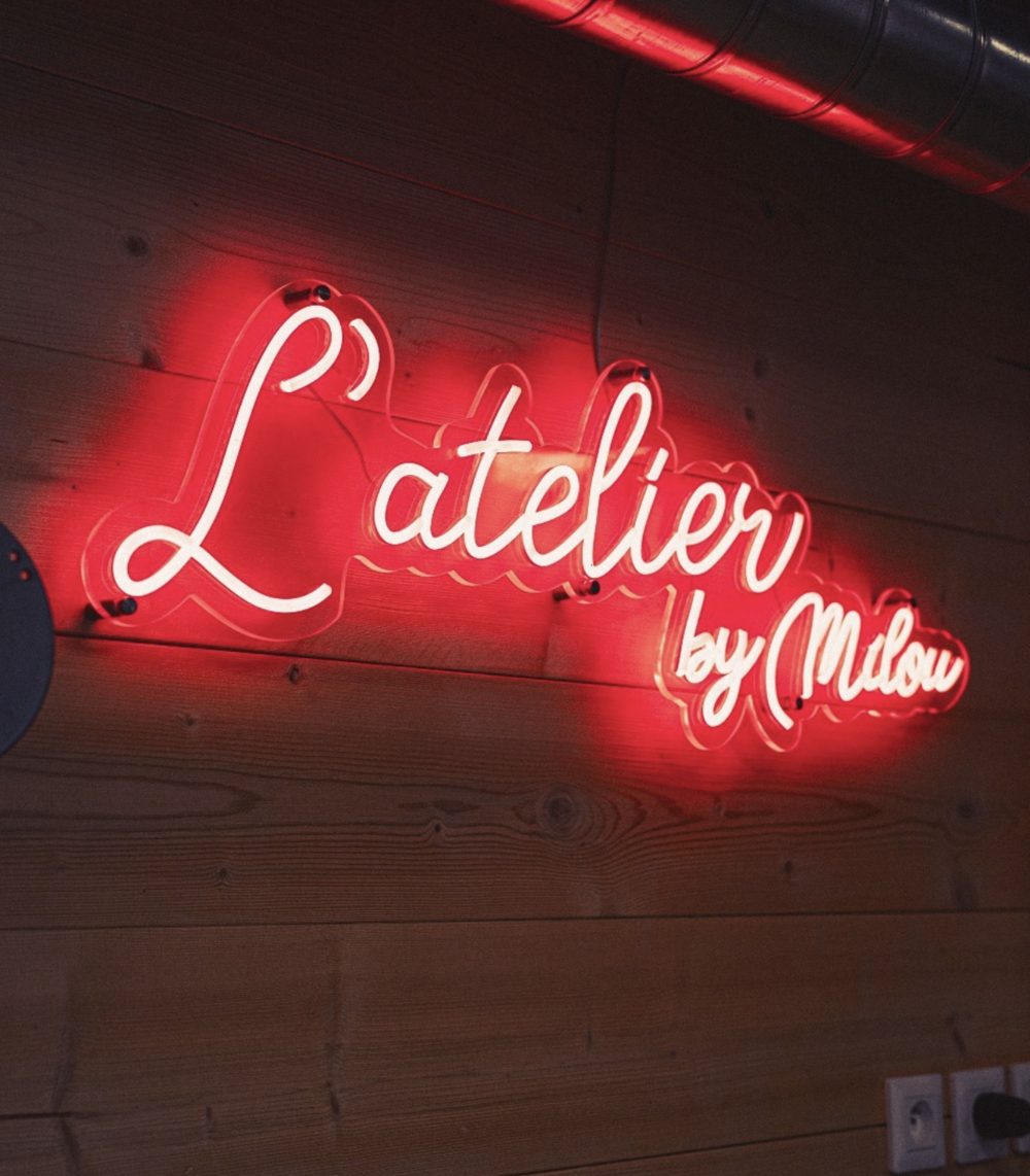 Neon sign “L'Atelier by Milou” in white letters with a red glow on a wooden plank wall
