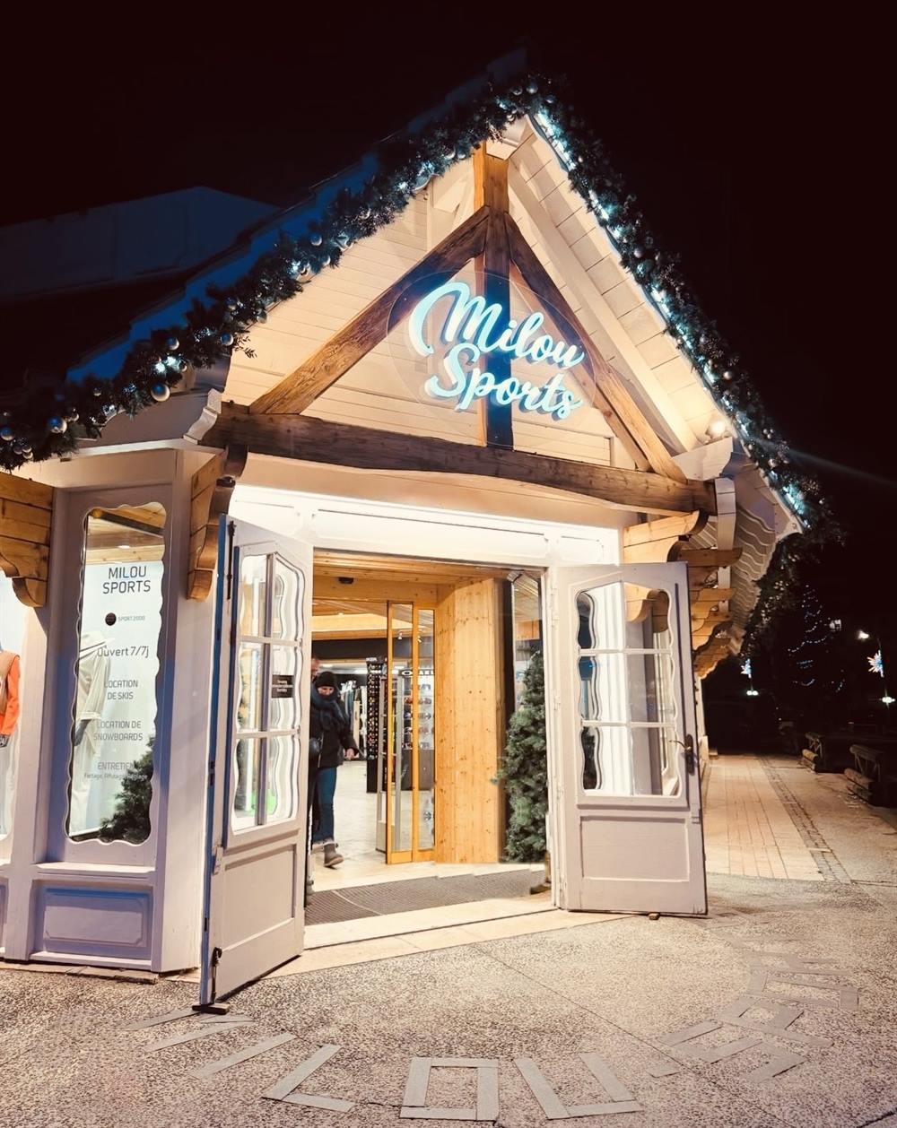 Het chalet van Milou Sports 's avonds verlicht, deuren wijd open naar de winkel, met de naam Milou in de bestrating