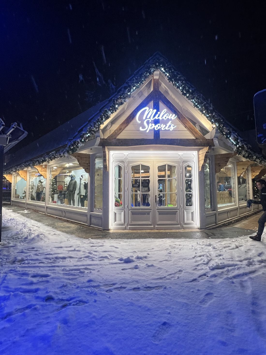 L'entrée du chalet Milou Sports illuminée la nuit sous la neige, avec sa porte blanche