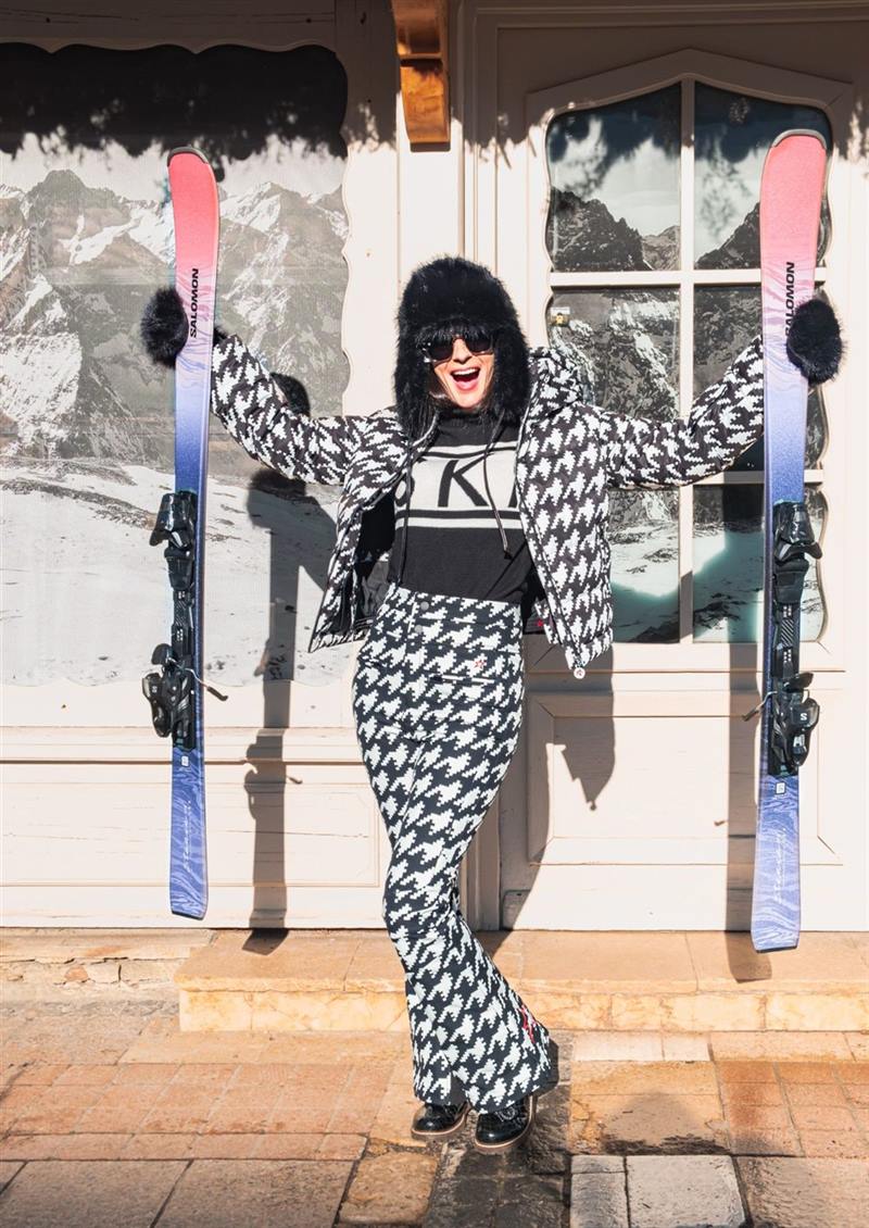 Lachende klant in een pied-de-poule skioutfit met in elke hand een Salomon-ski, voor de winkel van Milou Sports